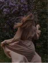 Woman in a flowing beige dress twirling near a garden with purple flowers.